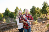 Family including mom and dad holding baby with christmas tree's behind them